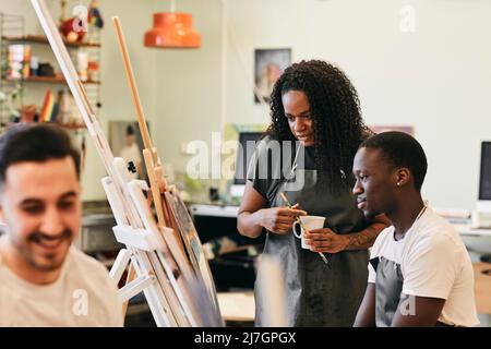 Female tutor looking at painting on easel by male student sitting in art class Stock Photo