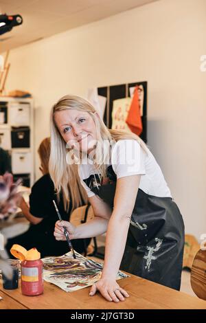 Portrait of smiling female mature student holding paintbrush at table in art class Stock Photo