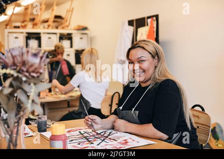 Portrait of smiling young female student painting on canvas while sitting at table in art class Stock Photo