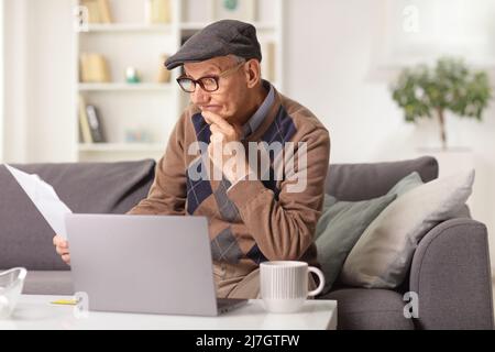 Elderly man sitting on a sofa at home and reading a paper document with a laptop computer on the table Stock Photo