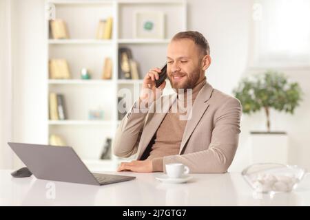 Happy young man sitting at home with a laptop computer on a table and making a phone call Stock Photo