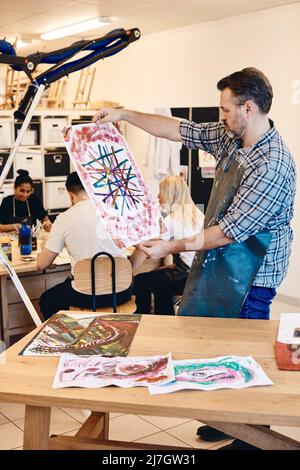Mature male instructor examining painting while students practicing in background at art class Stock Photo