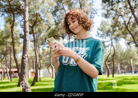 Young redhead woman wearing green tee with smiles looking at the phone. Sportive woman texting or scrolling or watching video on smart phone. Stock Photo