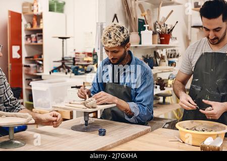Young man using work tool by friend molding clay while standing at table in art class Stock Photo