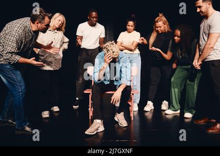 Distraught man scratching head while sitting amidst angry multiracial actors on stage Stock Photo