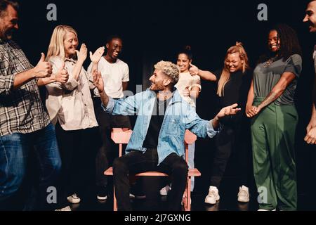 Happy young man sitting on chair amidst multiracial stage performers in class Stock Photo