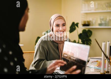 Smiling young woman in hijab sitting by freelancer with laptop at home Stock Photo