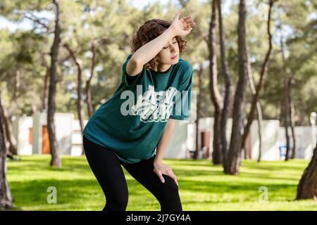 Young redhead woman wearing green tees and black yoga pants resting. Tired woman having rest after workout. Fitness model outdoors. Tired and exhauste Stock Photo