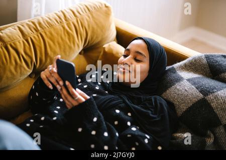 Smiling woman wearing hijab using mobile phone lying on sofa at home Stock Photo