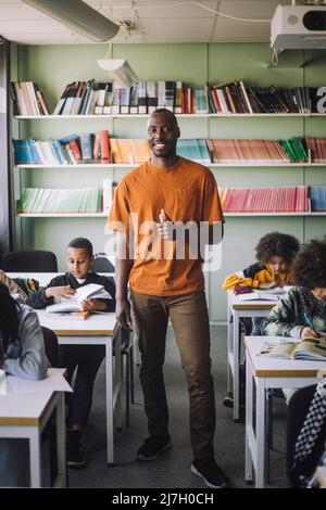 Portrait of confident teacher showing thumbs up while standing amidst students in classroom Stock Photo