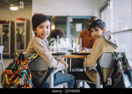 Portrait of schoolboys looking over shoulder while sitting on chair during lunch break Stock Photo