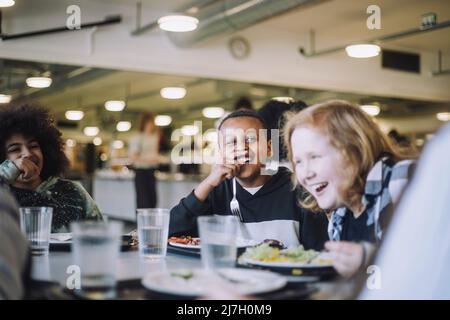 Friends laughing while having food at table during lunch break in cafeteria Stock Photo
