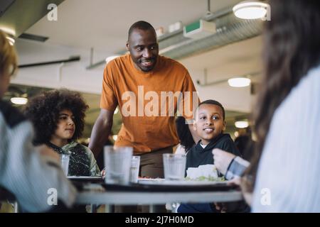 Smiling teacher standing by students during lunch break in cafeteria Stock Photo