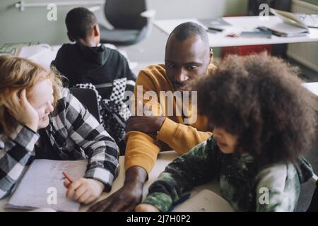 Male teacher interacting with pupils sitting at desk in classroom Stock Photo