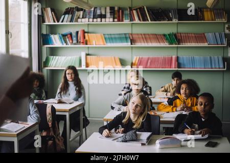 Diverse students writing while listening to teacher during lecture in classroom Stock Photo