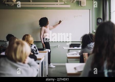 Side view of teacher explaining mathematics to students on white board in classroom Stock Photo
