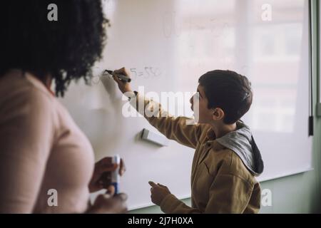 Side view of boy solving maths sum on white board with teacher in classroom Stock Photo