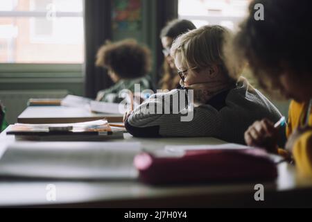 Side view of boy writing while leaning on elbow in classroom Stock Photo