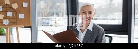 smiling banker with folder looking at camera near blurred corkboard on background, banner Stock Photo