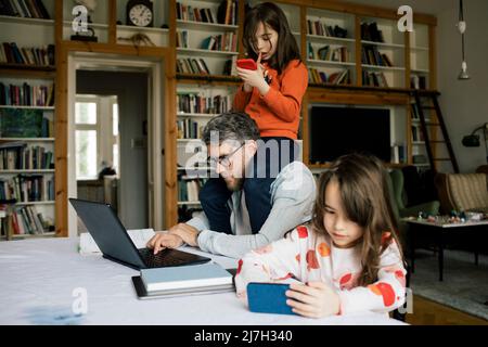 Daughter using smart phone by father working on laptop with girl sitting on shoulders at home Stock Photo