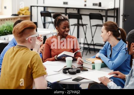 Cheerful group of young people studying together at round table in college library, focus on African American girl smiling Stock Photo