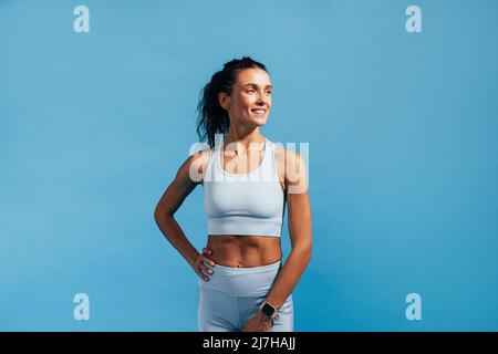 Smiling woman in fitness wear standing on blue background and looking away Stock Photo