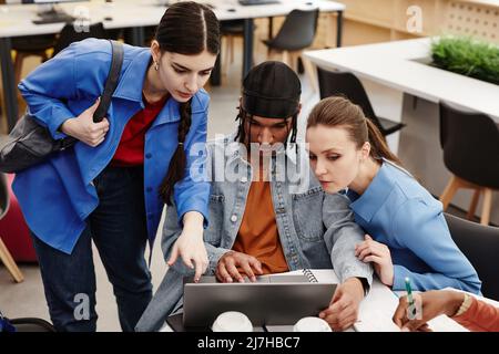 Diverse group of three college students using laptop together in library while working on project Stock Photo