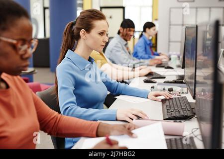 Vibrant side view shot of college students using computers in row, focus on Caucasian young woman doing research in library Stock Photo