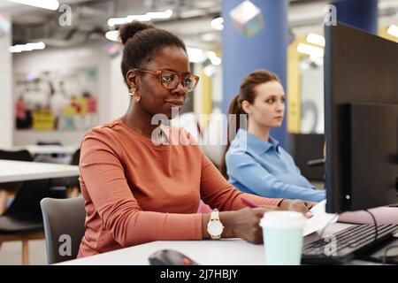 Vibrant portrait of young black woman using computer in college while doing research in library Stock Photo