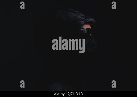 Stylish man in dark hoodie with black glasses and mask dark background, hacker, attacker and bandit Stock Photo