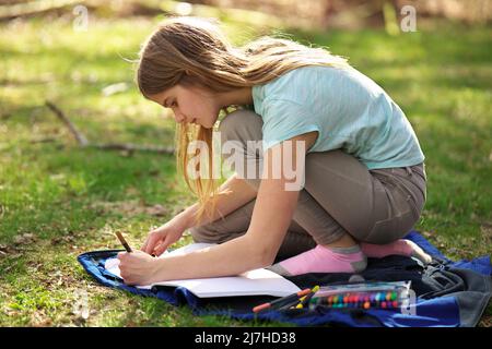 A young adolescent girl writes or colors in a notebook or journal on a blanket on the grass  Stock Photo