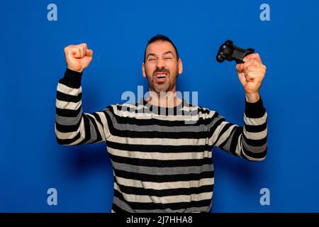 Photo of funny crazy amazed guy hold hand joystick playing video game, excited gamer came to finish first, raised fist, wear striped pullover Stock Photo