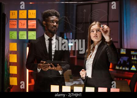 Team of young smart multiracial male and female office workers, standing near transparent glass board with colorful steakers and drawing graphs and charts for their new business project Stock Photo