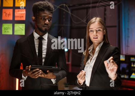 Team of young smart multiracial male and female office workers, standing near transparent glass board with colorful steakers and drawing graphs and charts for their new business project Stock Photo