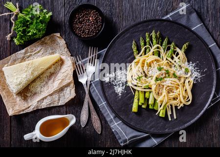 roasted asparagus spears with spaghetti sprinkled with shredded pecorino cheese and parsley on black plate on dark wooden table with ingredients, hori Stock Photo