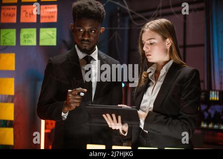 Night work at office, project deadline. Male and female multiethnic creative colleagues, using digital tablet and discuss project with sticky notes on a glass wall. Stock Photo