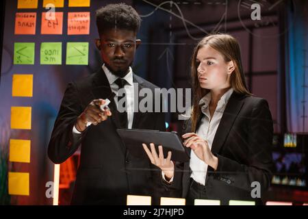 Night work at office, project deadline. Male and female multiethnic creative colleagues, using digital tablet and discuss project with sticky notes on a glass wall. Stock Photo
