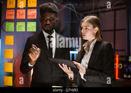 Technology, software programming, idea, people concept. Waist up portrait of two young diverse multiethnic coworkers, standing near the glass board, writing and using tablet pc Stock Photo