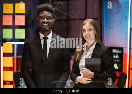 Happy mixed race young colleagues posing behind glass kanban scrum board with business project working process. Smiling employees using management tools for work planning. Stock Photo