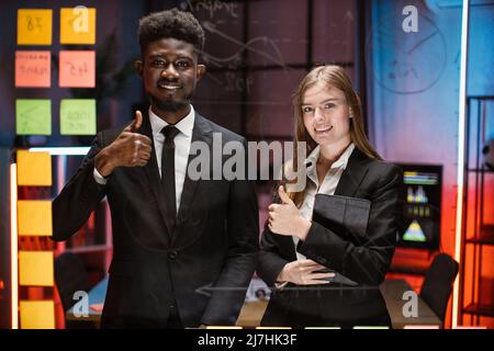 Happy mixed race young colleagues posing behind glass kanban scrum board with business project working process. Smiling employees using management tools for work planning showing thumb up. Stock Photo