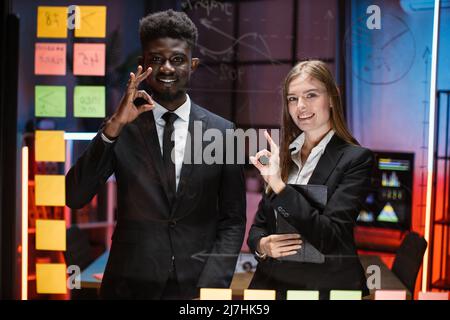 Happy mixed race young colleagues posing behind glass kanban scrum board with business project working process. Smiling employees using management tools for work planning showing sign ok. Stock Photo