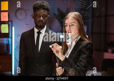 Team of two professional multicultural traders discussing possible risks and profits on trade market. Male and female partners looking at glass board while standing in modern office conference room. Stock Photo