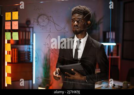 Close up of African American businessman, working on project plan using sticky papers on glass wall, scrum board. Confident executive manager, holding digital tablet Stock Photo