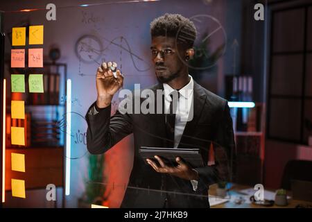 Handsome smart African business man, writing ideas on glass board, working in the night at office and using digital tablet. Male ceo analyzing financial charts and kanban stickers on the board Stock Photo