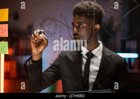 Close up blurred portrait of young handsome African American man, writing on glass board and making project plan analysis. Economics, trade business, finance and corporate concept Stock Photo