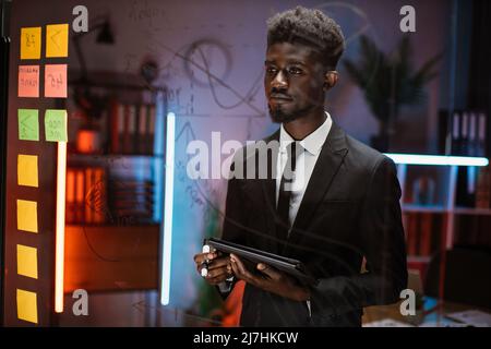 Focused IT specialist project manager standing near big glass board at office and looking at set of stickers and graphs, planning the meeting. African American man with tablet pc works in the evening Stock Photo