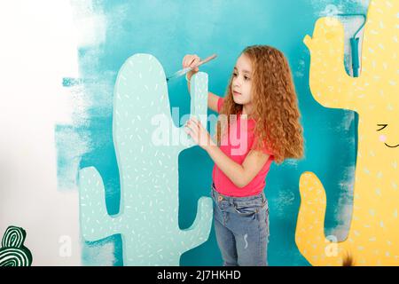 adorable little girl is doing repairs in the apartment fun paints the wall. Portrait of a beautiful smiling child with a paint roller and big paper ca Stock Photo