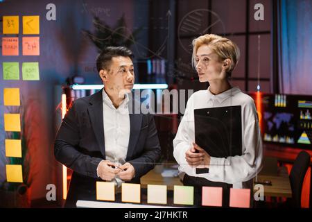 Confident blond mature businesswoman mentor explaining business strategy and tasks on stickers on glass board to focused male Asian coworker. Diverse employees working together on new startup Stock Photo