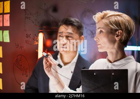 Close up view through glass wall of asian and caucasian colleagues working together in office, searching solution of problem, writing on board with sticky notes, analyzing financial charts Stock Photo