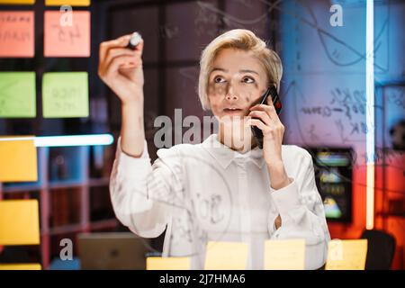 Strategy, business and scrum kanban concept. Confident experienced Caucasian female manager or ceo holding marker and writing on glass board in office room, while talking with colleague on phone Stock Photo